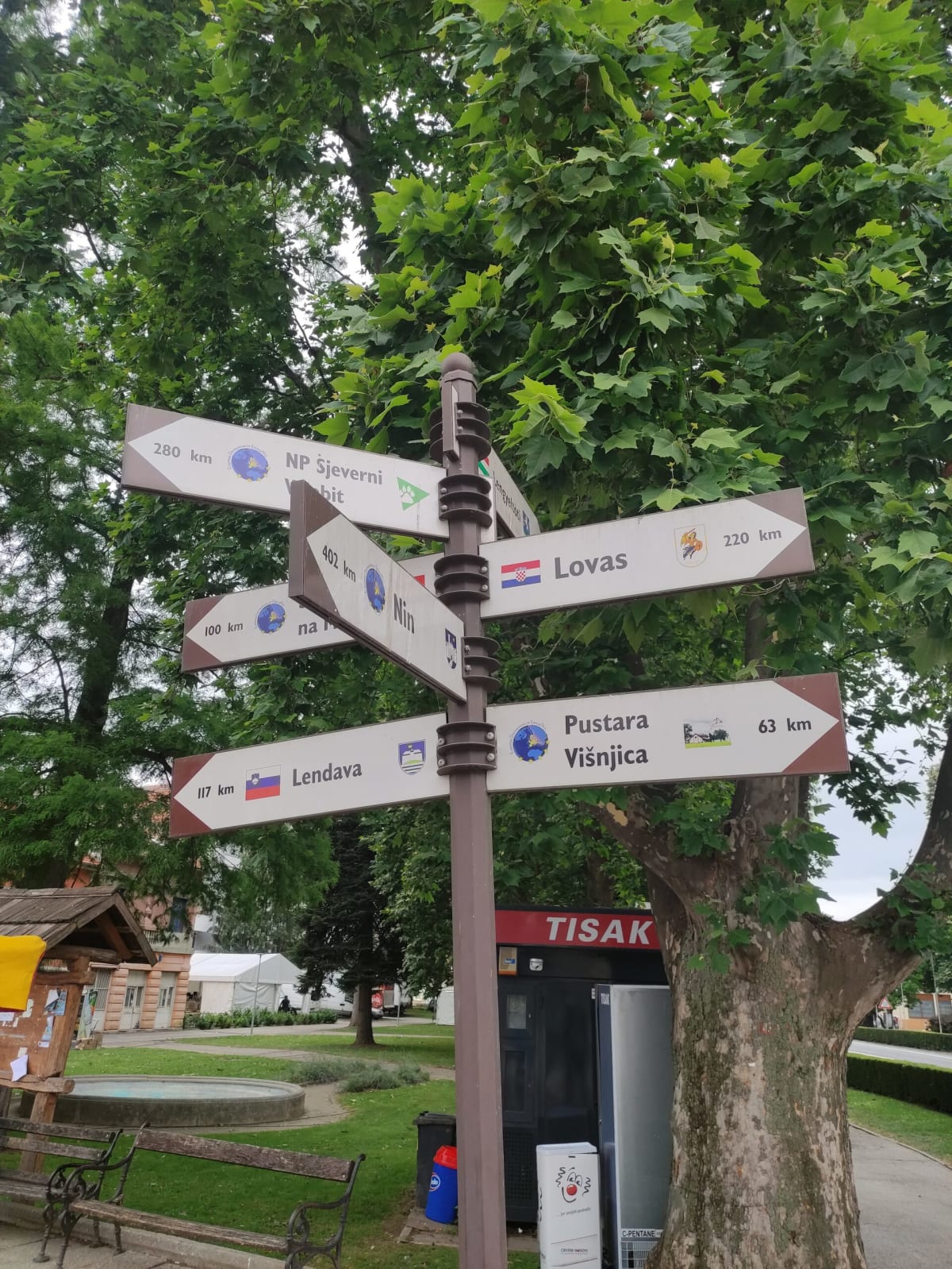 A multi-armed tourist signpost in the park pointing to partner towns with distances — Lendava, Nin, Lovas, Pustara Višnjica — a plane tree and a Tisak kiosk behind