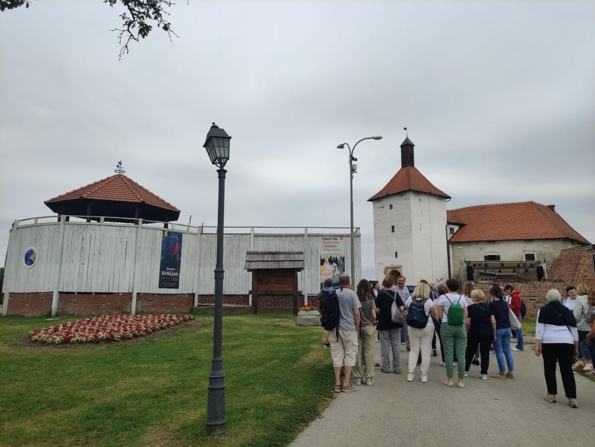 The Stari grad fortress of Đurđevac — a whitewashed defensive tower and red-roofed wings behind a wooden palisade, a banner for the Toulouse-Lautrec exhibition, a tour group gathered at the entrance