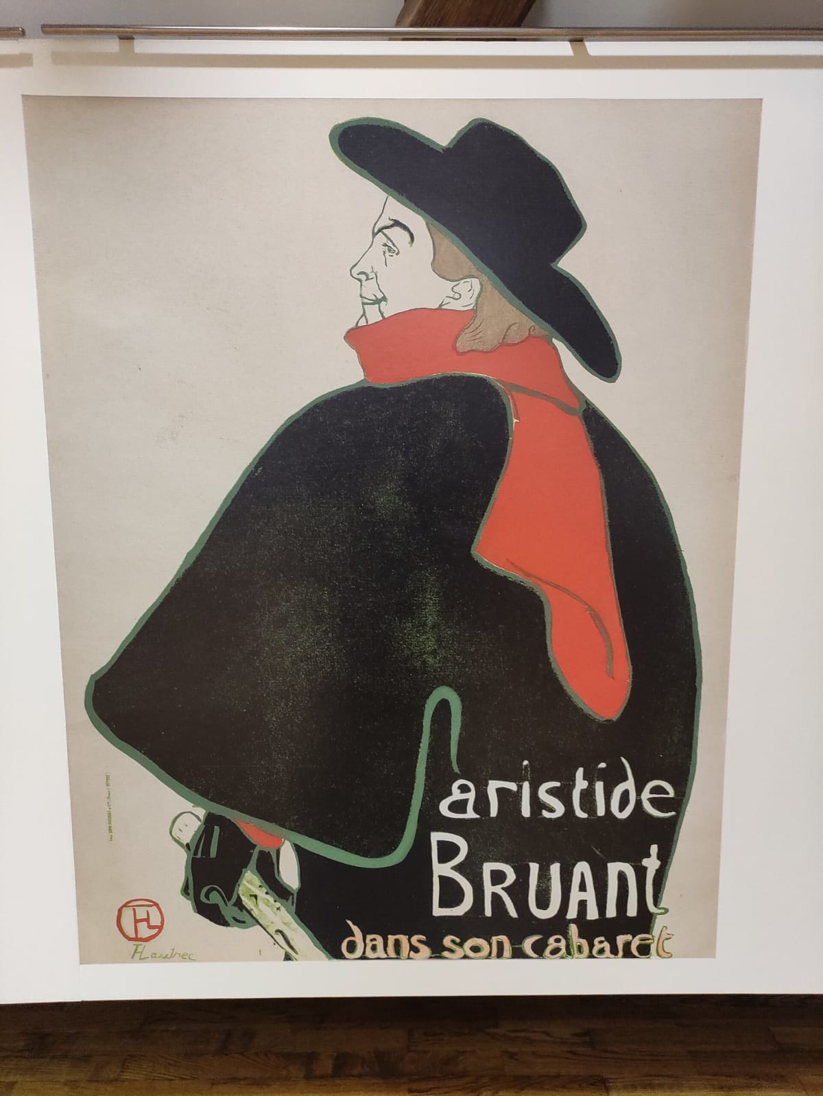 Toulouse-Lautrec's poster "Aristide Bruant dans son cabaret" on the wall — the singer in profile in a black cloak, red scarf and broad black hat against a pale ground