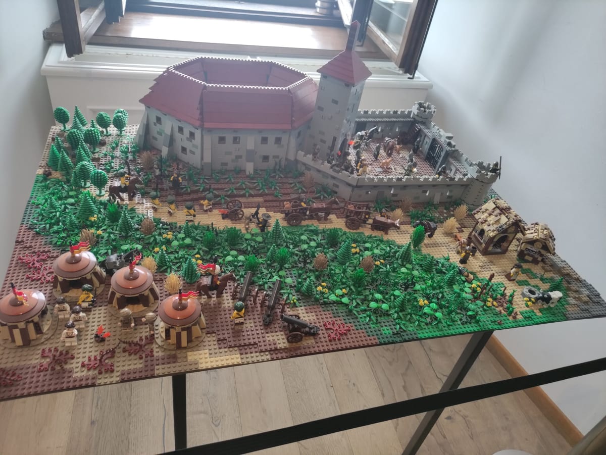 A large LEGO model of the Đurđevac fortress and its siege, with minifigure soldiers, cannon, tents and trees, displayed on a table by a window