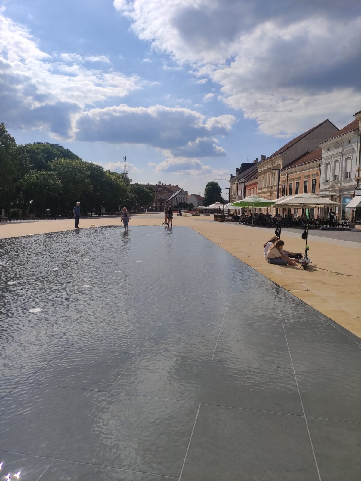 Koprivnica's main square in the afternoon — a long shallow black reflecting pool set into the paving, café umbrellas and 19th-century townhouses down one side, people wading and sitting on the edge
