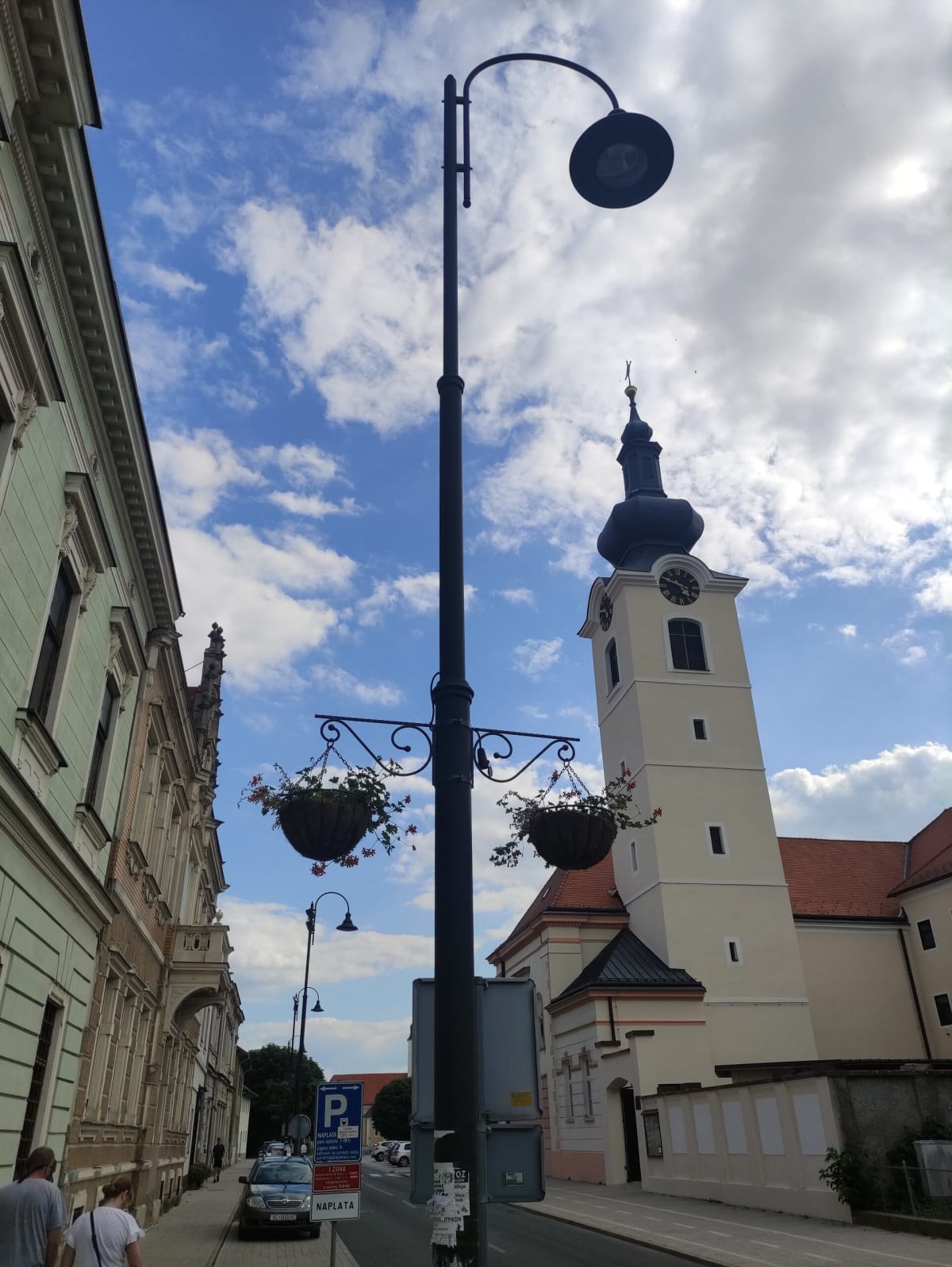 A Koprivnica street looking up past a tall wrought-iron lamp-post with hanging flower baskets to the onion-domed Baroque bell tower of the parish church