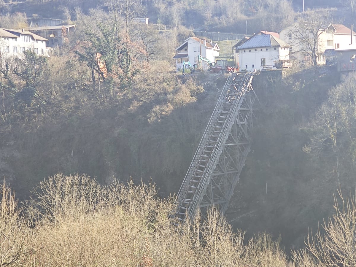 The twisted remains of the Jablanica railway bridge hanging down into the gorge, houses and an excavator on the rim above, bare winter scrub in the foreground