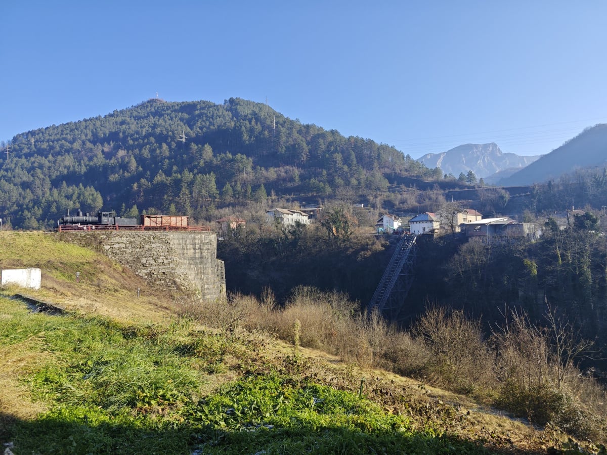 The locomotive on its stone plinth at left and the broken bridge across the gorge at right, forested hill and snowy peaks under a clear blue sky