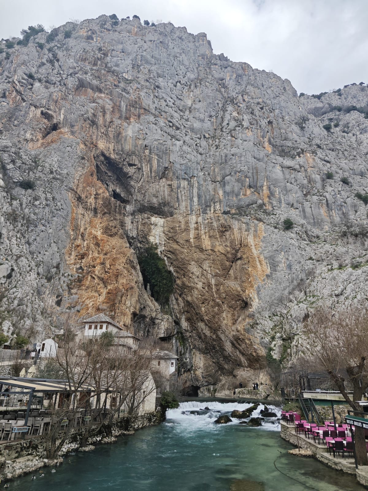 The Buna spring at Blagaj — turquoise water rushing from beneath a huge orange-and-grey limestone cliff, riverside restaurants with pink chairs on either bank
