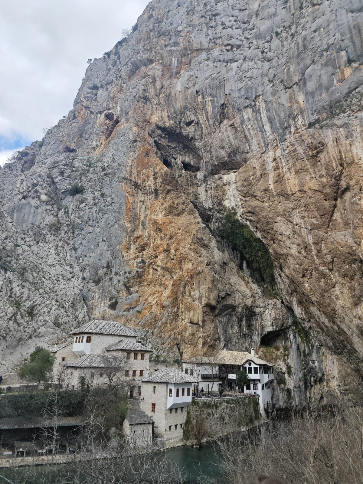 The white stone tekke of Blagaj tucked at the foot of a sheer striped cliff above the Buna, a dark cave mouth beside it