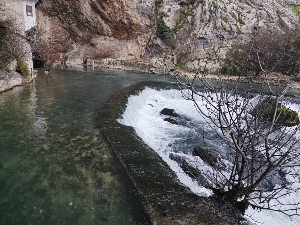 A small weir on the Buna below the cliff, white water pouring over rocks beside a bare winter tree and clear green shallows