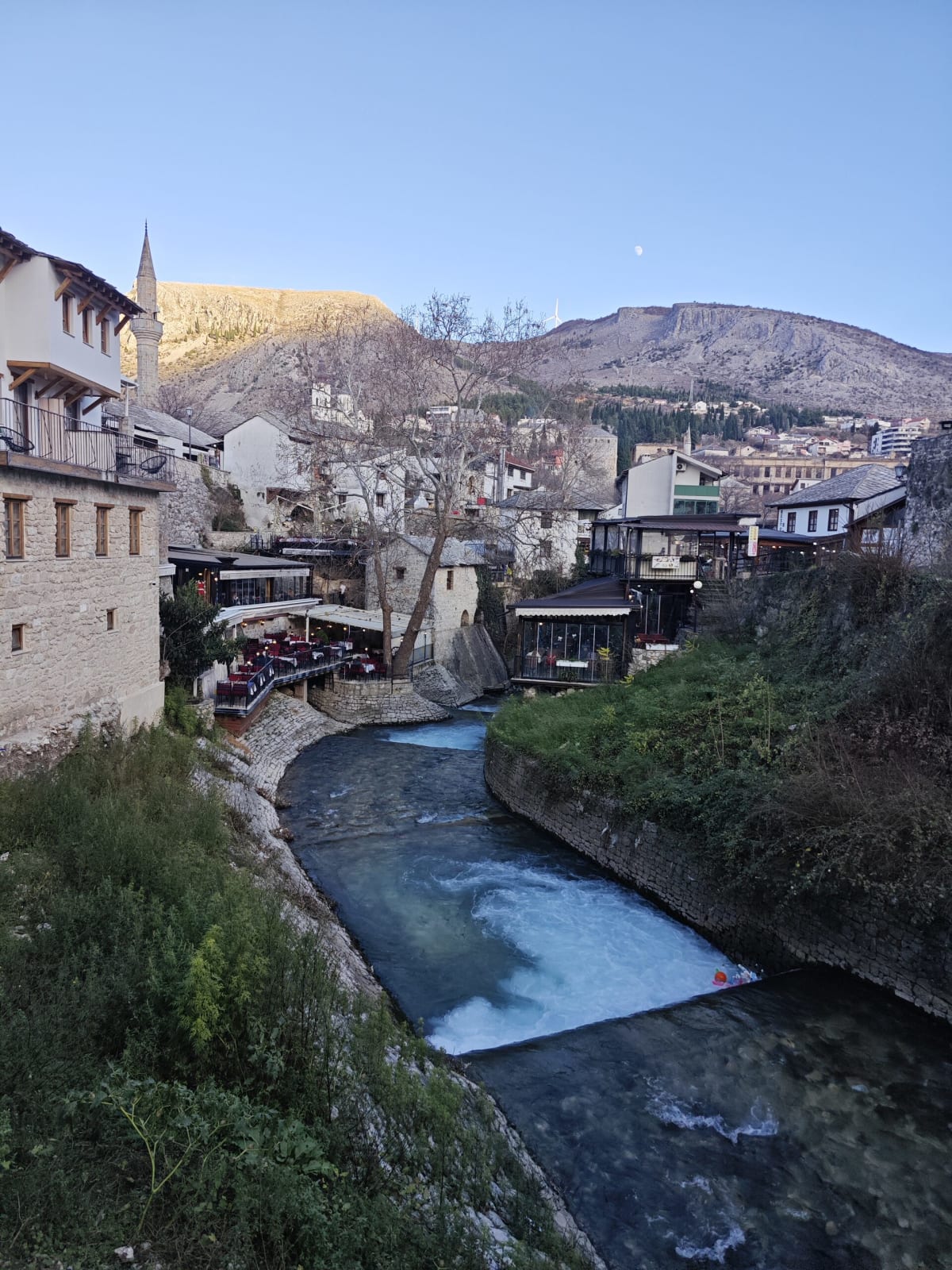 The Kriva Ćuprija stream cutting through Mostar's old town — stone houses, terraced restaurants and a minaret, with bare hills and a pale moon above