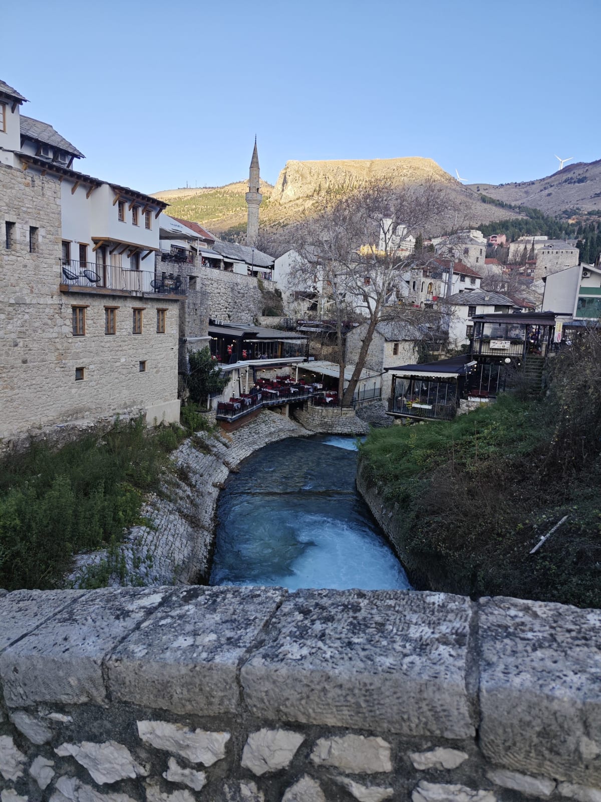 The same stream from the edge of the stone bridge, the old town's balconied stone houses and minaret rising above the blue-green water