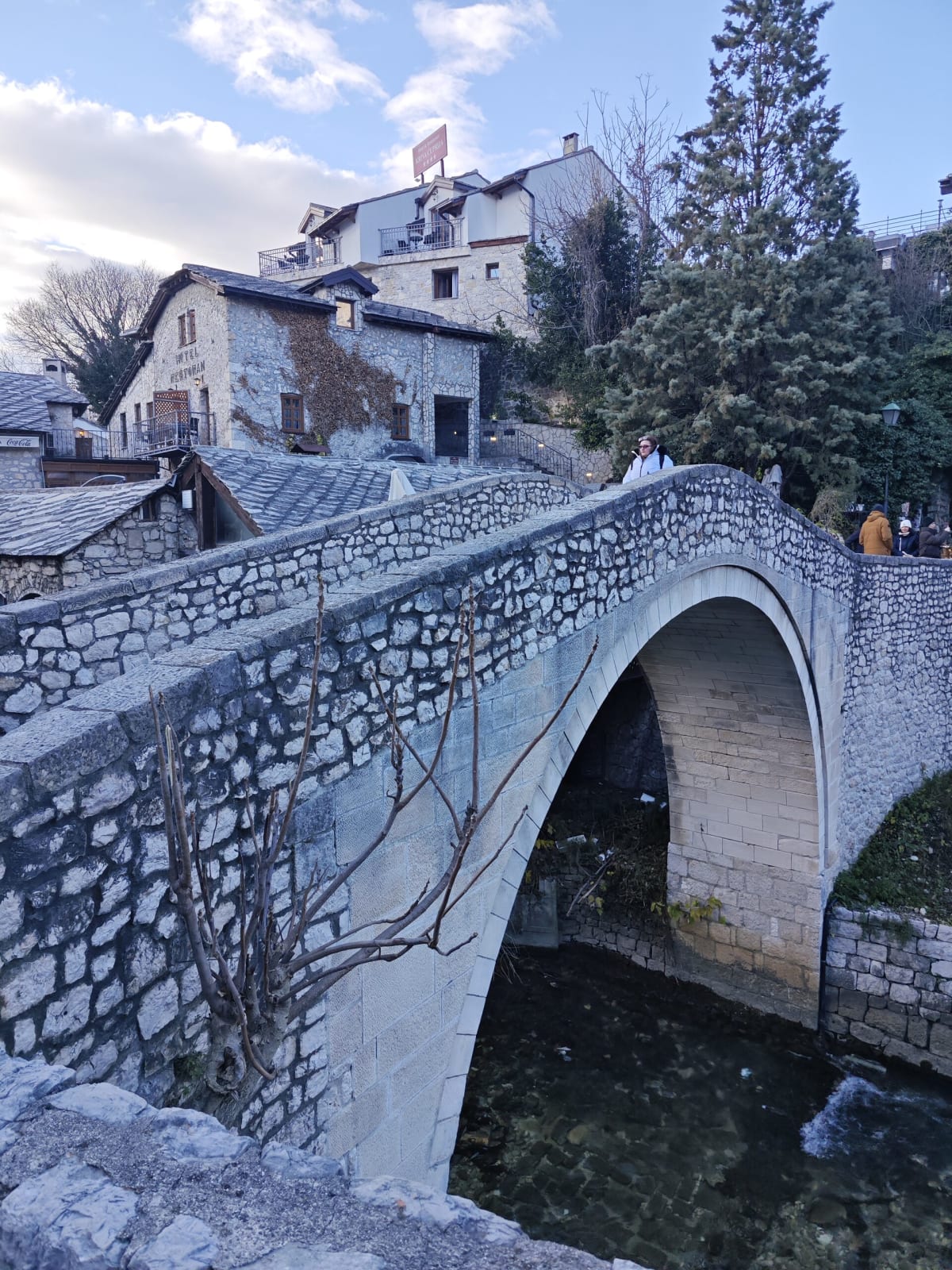 The Crooked Bridge, a small humpbacked stone arch, with slate-roofed houses behind and pine trees against a cloud-streaked sky