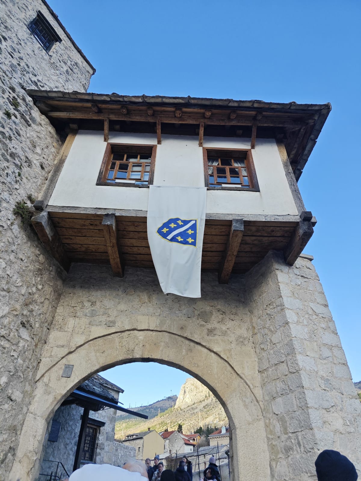 Looking up at the Halebija tower gate at the end of Mostar's Old Bridge — a timber-framed upper storey with a Bosnian lily flag hanging below it