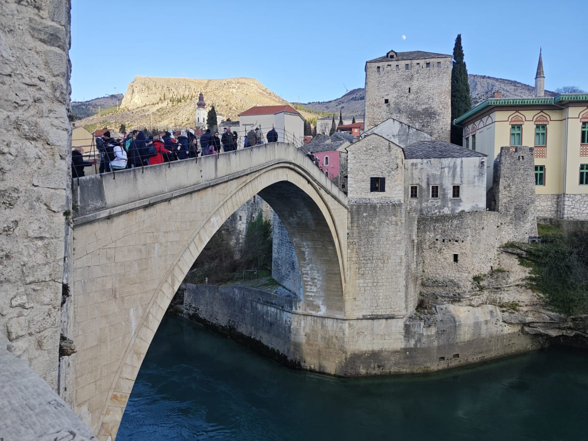 The Old Bridge in Mostar seen from the east bank, crowds along its crest, the Tara tower and a green-roofed mosque beyond, a thin moon in the blue sky