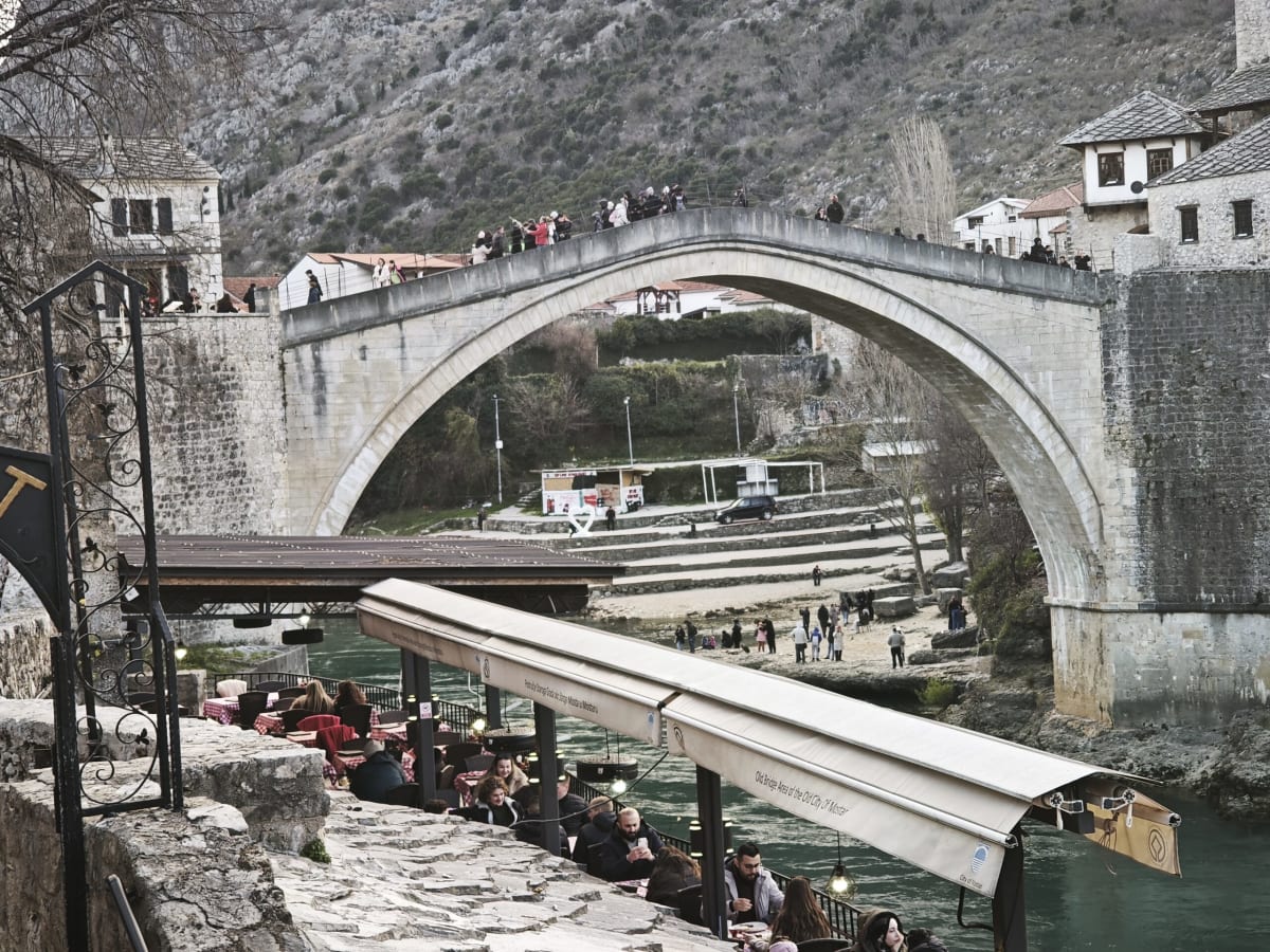 The arch of the Old Bridge framed from a riverside café terrace, diners under awnings and the steep dry hillside behind