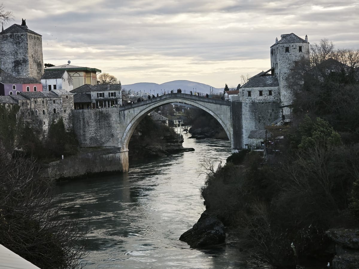 Mostar's Old Bridge at dusk from upstream, the Neretva sweeping toward it between dark banks and stone towers under a pale cloudy sky