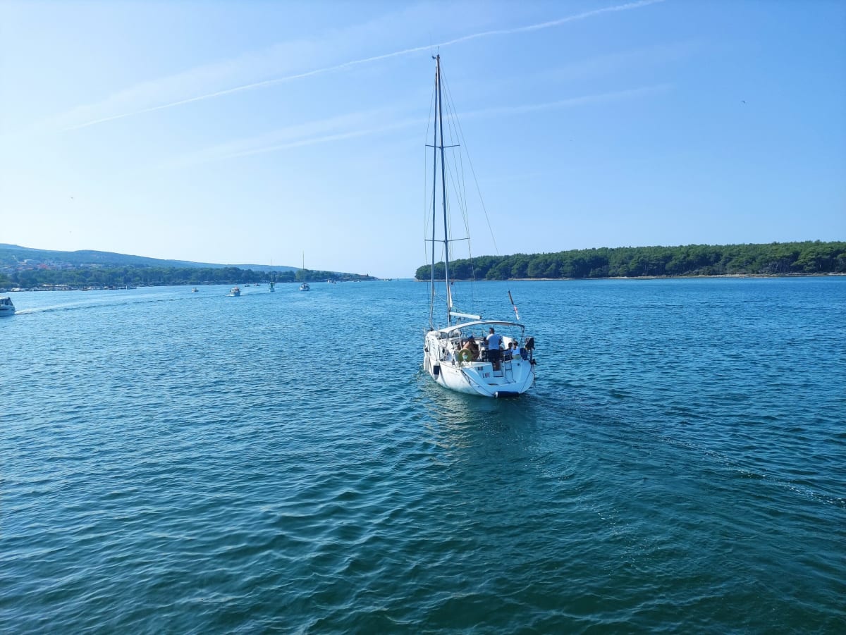 A chartered sailing yacht named Lady motoring across the calm bay past the pine-covered shore, other small boats and aircraft contrails above