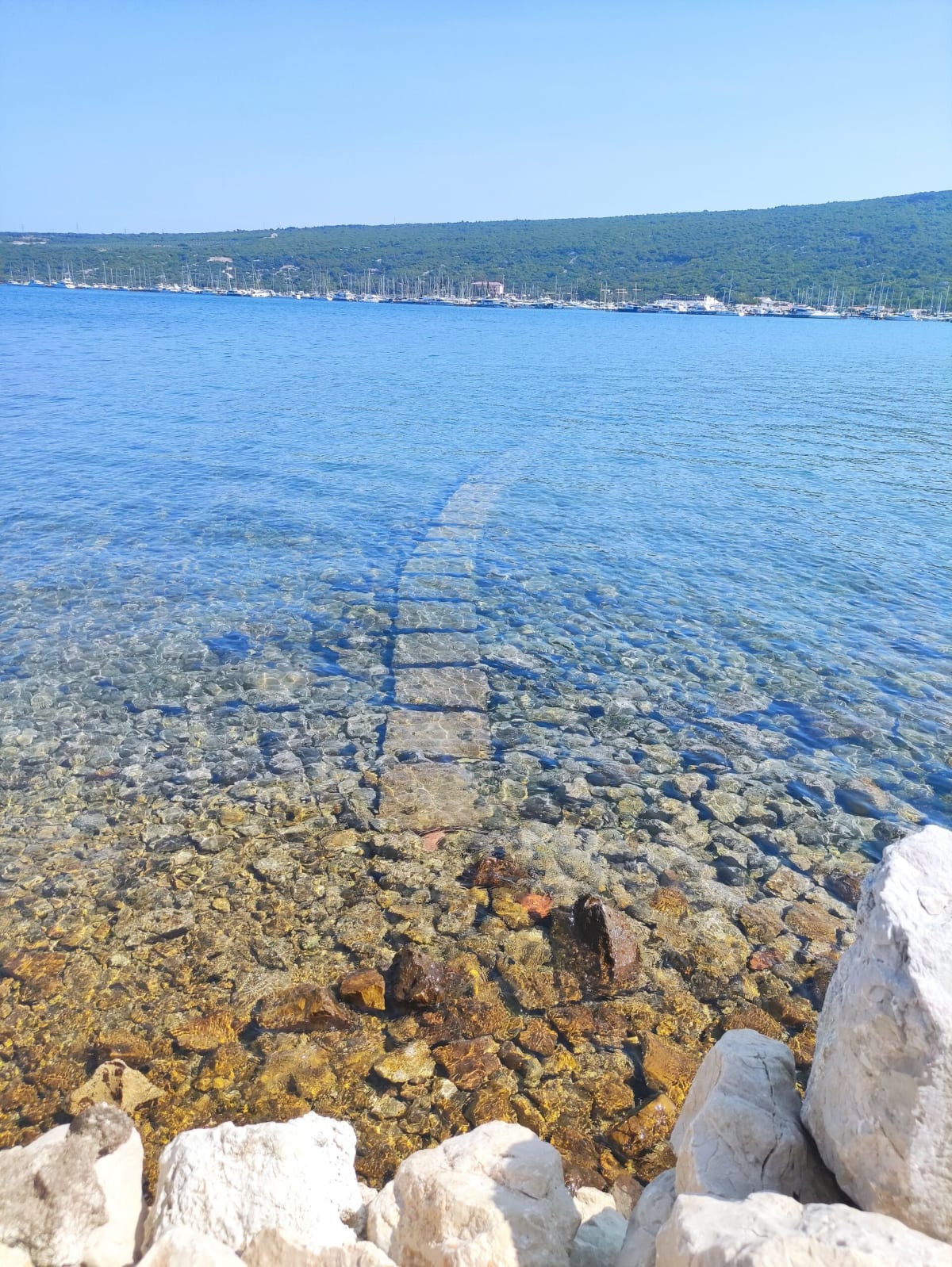 A submerged stone path running out from the shore across a shallow, crystal-clear seabed, Punat's mast-filled marina on the far side of the bay