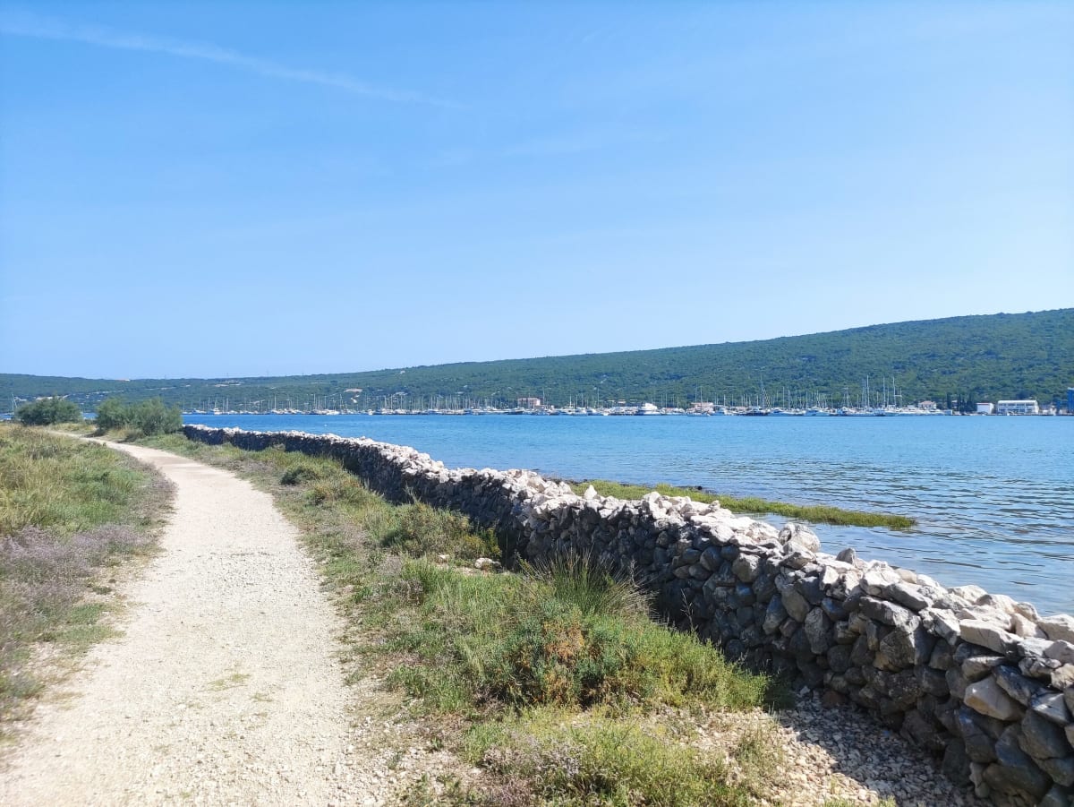 A gravel path along a low drystone wall on the Košljun shore, the still bay and Punat marina beyond