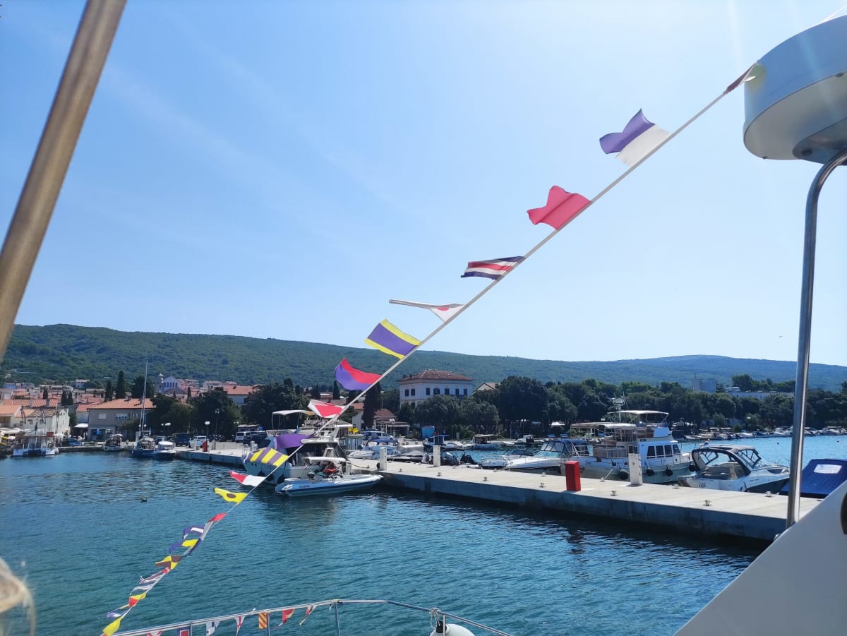 Leaving Punat by boat under a string of bright signal flags, the palm-lined waterfront and an ochre mansion along the quay
