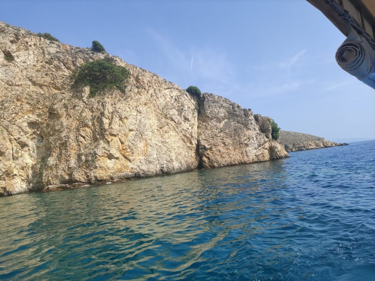 Pale, fractured limestone sea-cliffs dropping straight into deep blue-green water, shot from a boat rounding the headland