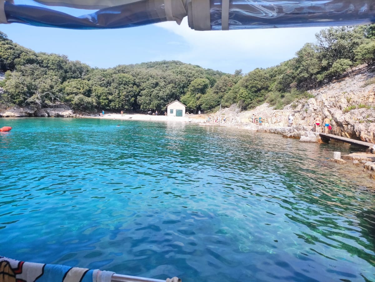 A sheltered cove with a small pebble beach and a stone boat-house backed by oak woods, swimmers in the turquoise shallows