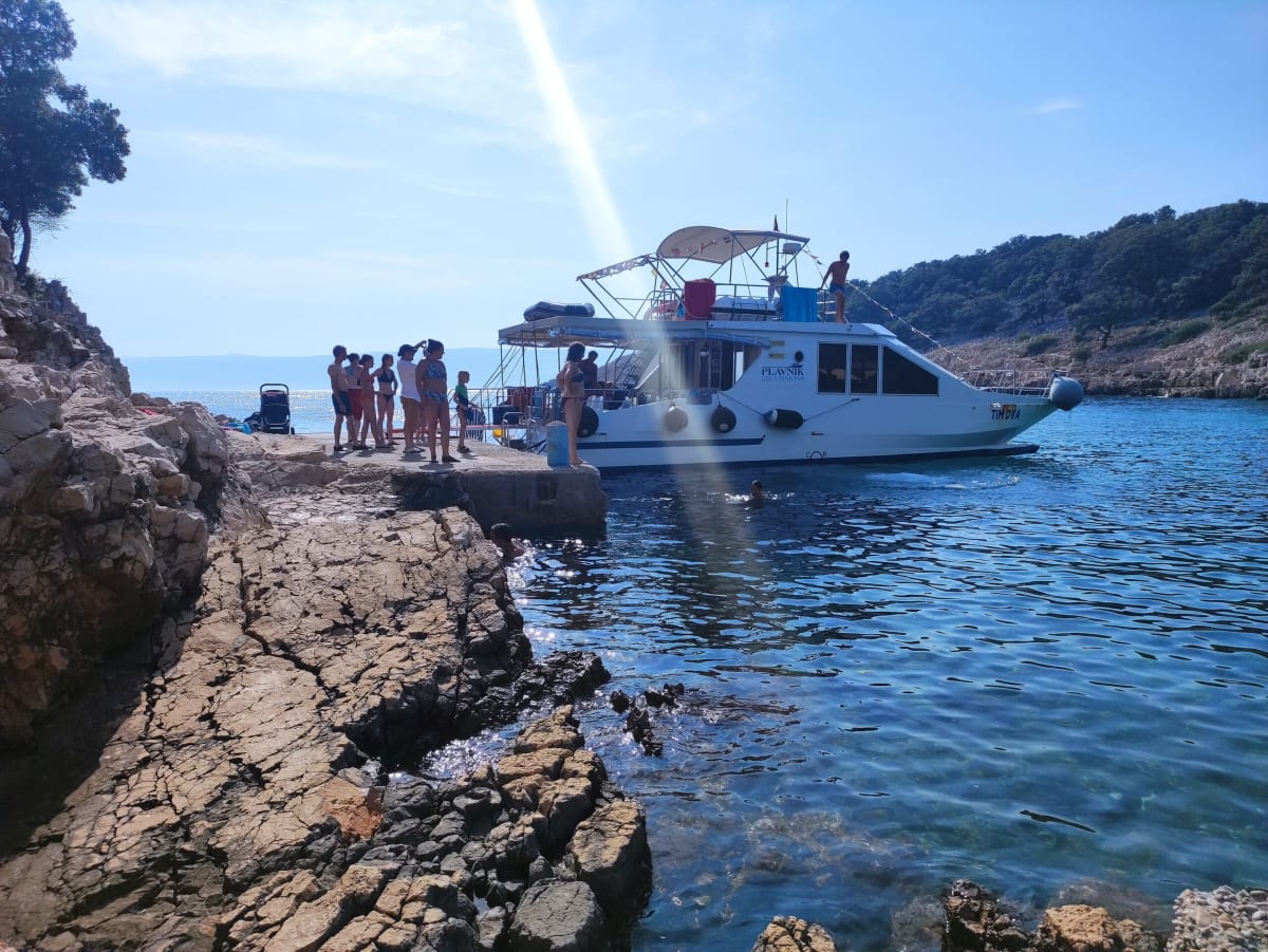 The excursion boat Tim Dva moored against a rocky quay in the cove, a group of passengers standing on the rocks and a child swimming alongside