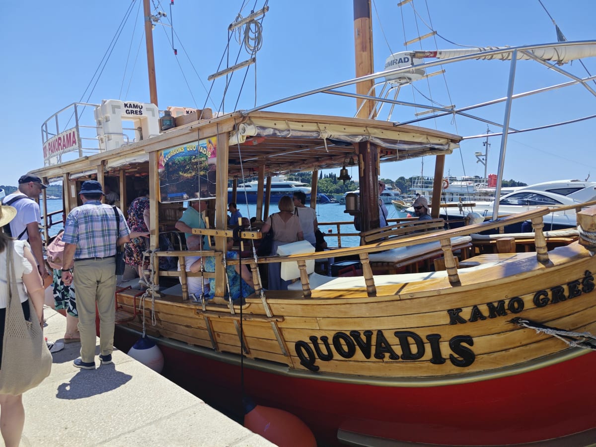 Passengers boarding the wooden excursion boat Quo Vadis at the quay, its varnished deck, red hull and twin masts lit by strong midday sun