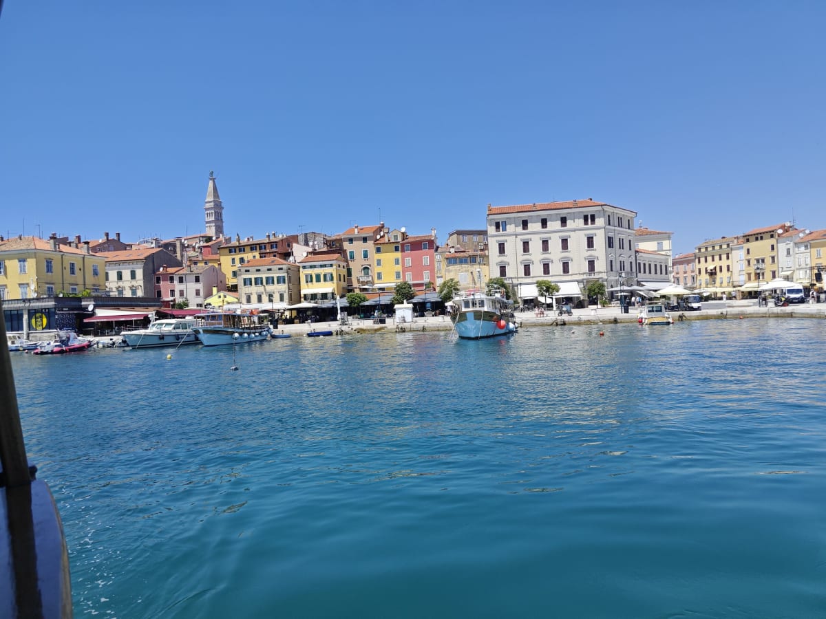 Rovinj's colourful waterfront from the water — pastel façades along the quay beneath the bell tower, small boats moored in front