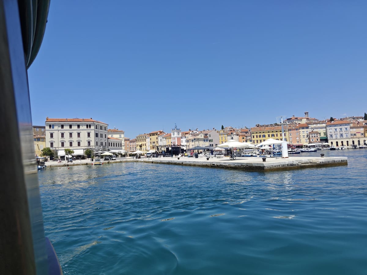 The Rovinj harbour front from a boat, a big pale palazzo and café umbrellas along the quay across clear blue-green water