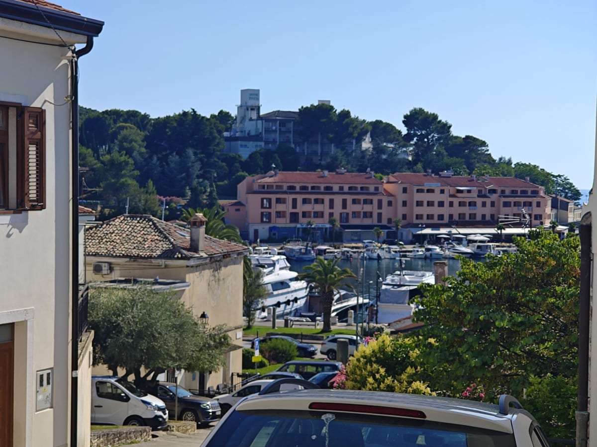 A small harbour seen from behind a parked car — a pink hotel building along the quay, yachts moored in front and pines on the hill above