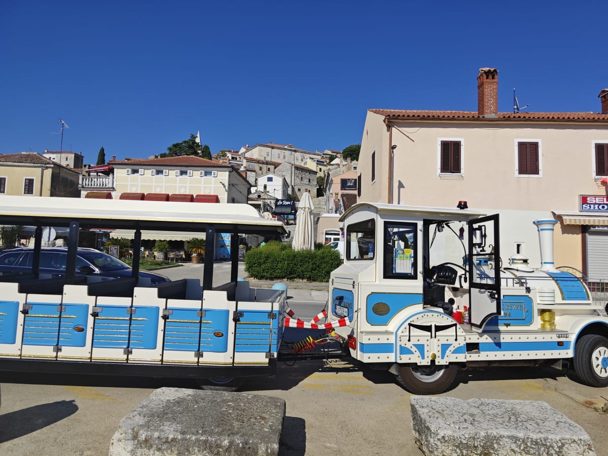 A blue-and-white tourist road-train parked in a sunlit square beside old stone houses and a terracotta-roofed town above