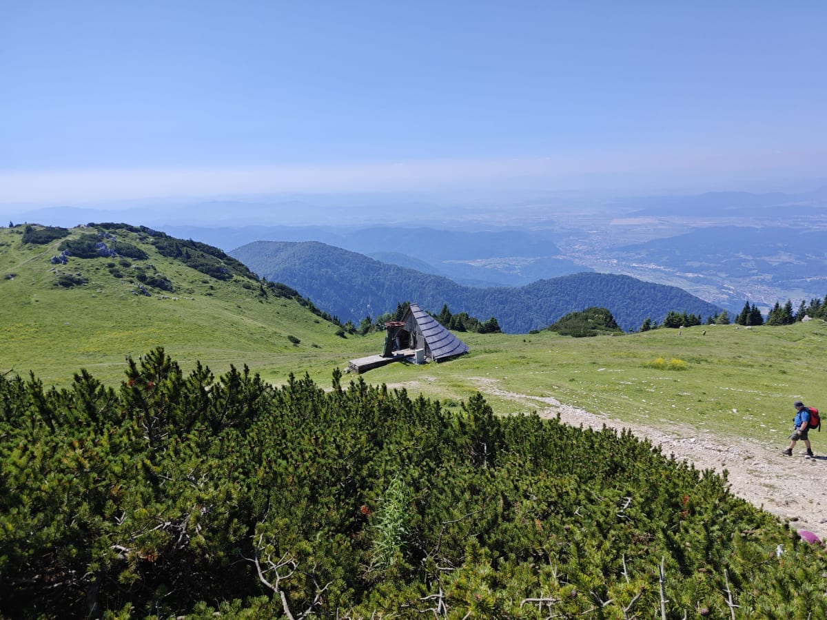 A high green plateau on Velika Planina with a small chapel in the distance, a hiker on the path and the valley and ridges hazy far below