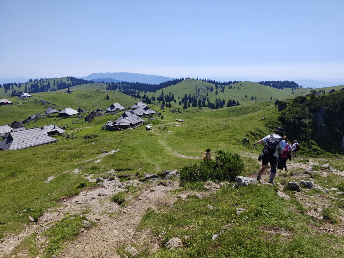 Hikers on a rocky path above the grassy basin of Velika Planina, clusters of shingle-roofed herdsmen's huts dotting the hills