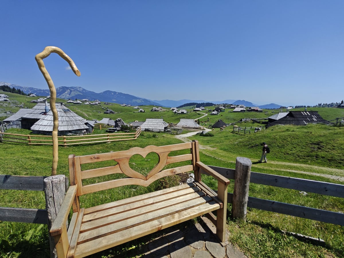 A wooden bench with a carved heart and a crooked staff beside it, looking out over the huts of Velika Planina and a ridge of mountains