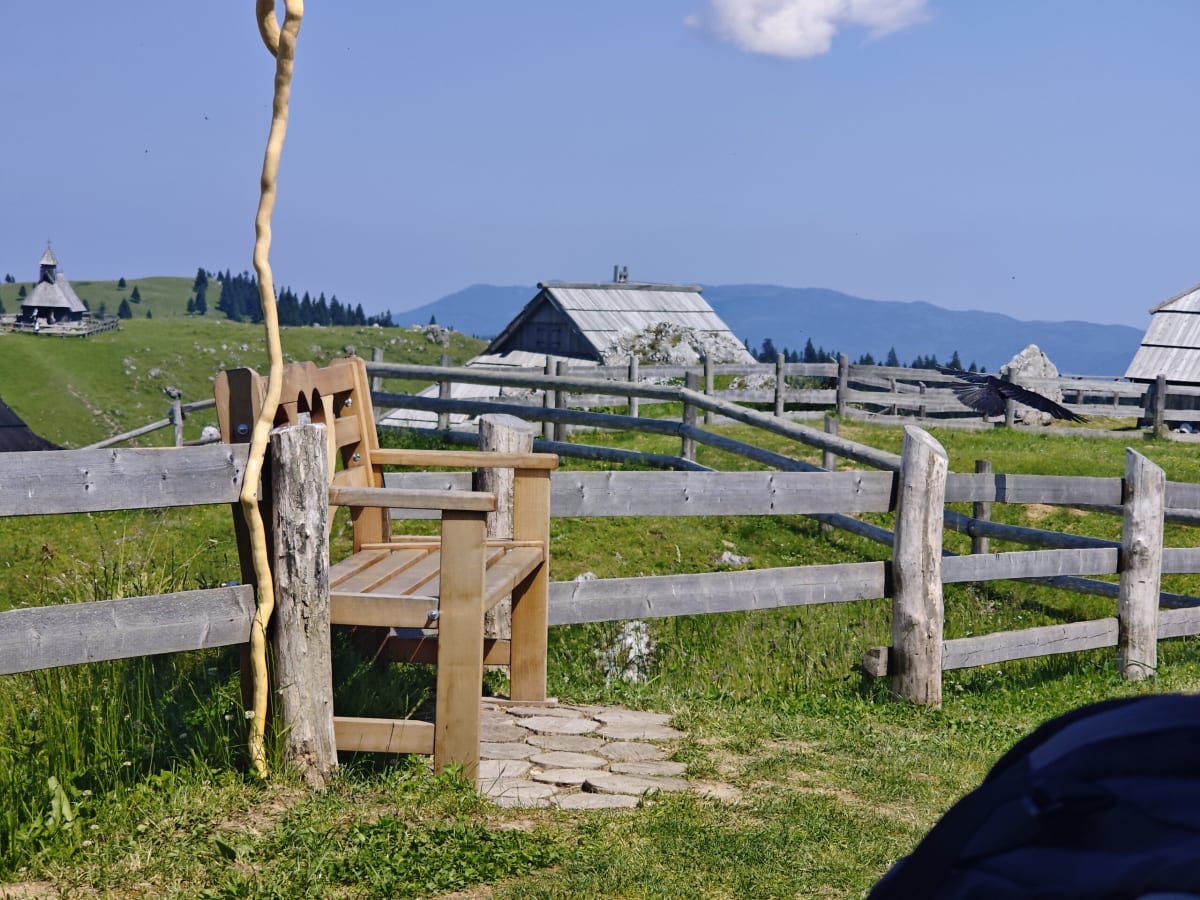 A wooden armchair-bench on a stone slab behind a log fence, a twisted staff beside it, huts and a chapel on the ridge behind