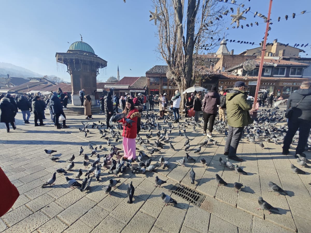 The Sebilj, Sarajevo's green-domed wooden fountain, on a sunlit Baščaršija square thick with pigeons and people, a girl in a red jacket feeding them in the foreground