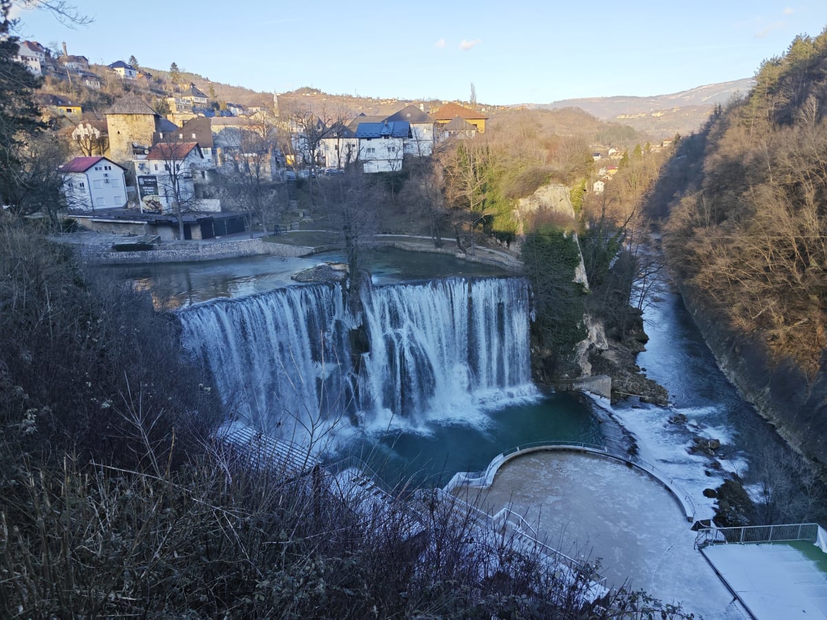 The Pliva waterfall in Jajce seen from the hillside — a wide curtain of white water dropping into a turquoise pool, old houses and a stone tower on the bank above, ice on the paths at lower right
