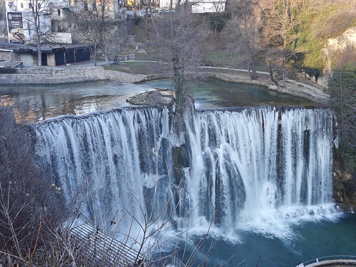 A closer view of the Jajce waterfall, two broad cascades split by a rock with a bare tree, the river calm above and winter woods behind