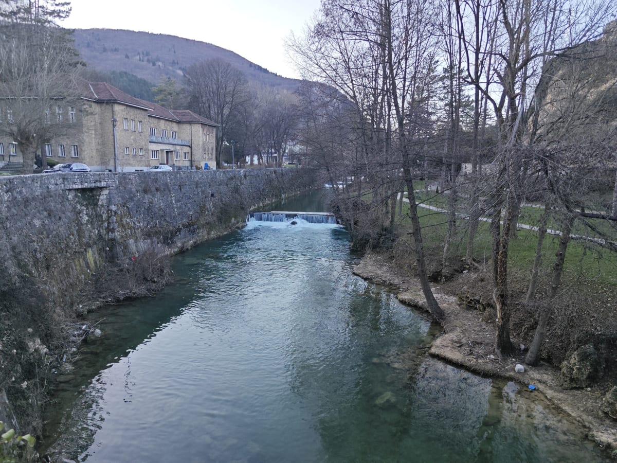 The Pliva river in Jajce below the falls, green water running between a high stone wall and a bare-treed bank, a small weir ahead and a pale building at left