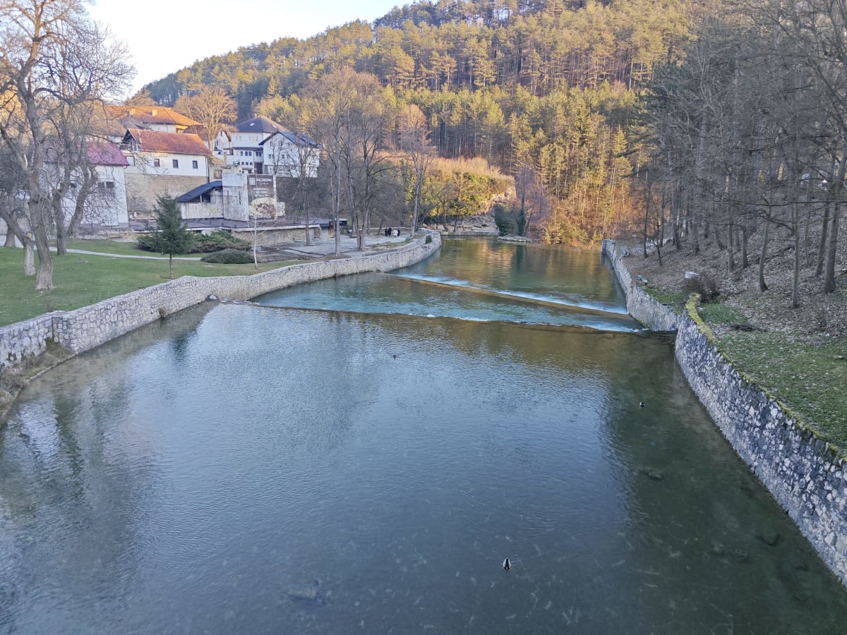 The river bend in Jajce lined with low stone embankments, a small weir across the water, pine-covered hills and red-roofed houses in late golden light