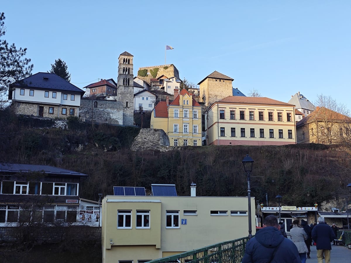 Jajce's old town stacked up the hillside — the slender Church of St Luke's bell tower, a ruined fortress wall and yellow and white houses under a clear sky