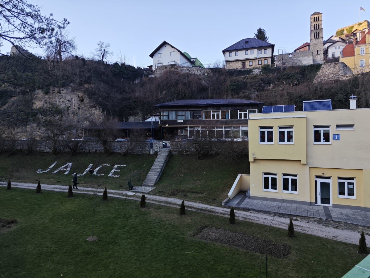 JAJCE spelled out in white stones on a lawn below the old town, the bell tower and townhouses on the slope above and a yellow building with solar panels at right