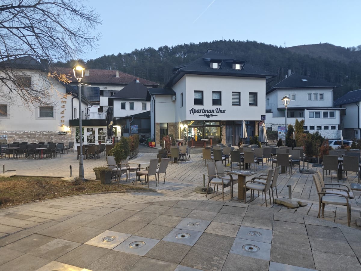A small Jajce square at dusk — café terraces with tables and chairs, street lamps just lit, a white guesthouse and wooded hills behind
