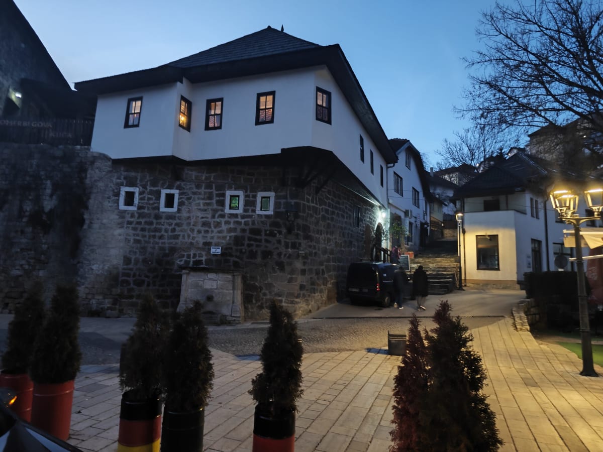 A white upper-storey house with a jutting timber-framed front above a dark stone wall, lit windows glowing as evening falls over a stepped lane