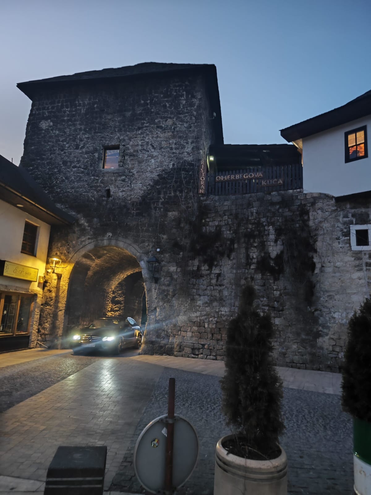 The old stone gate tower of Jajce at dusk, a car emerging with its headlights on from the vaulted passage, a restaurant sign above the wall at right