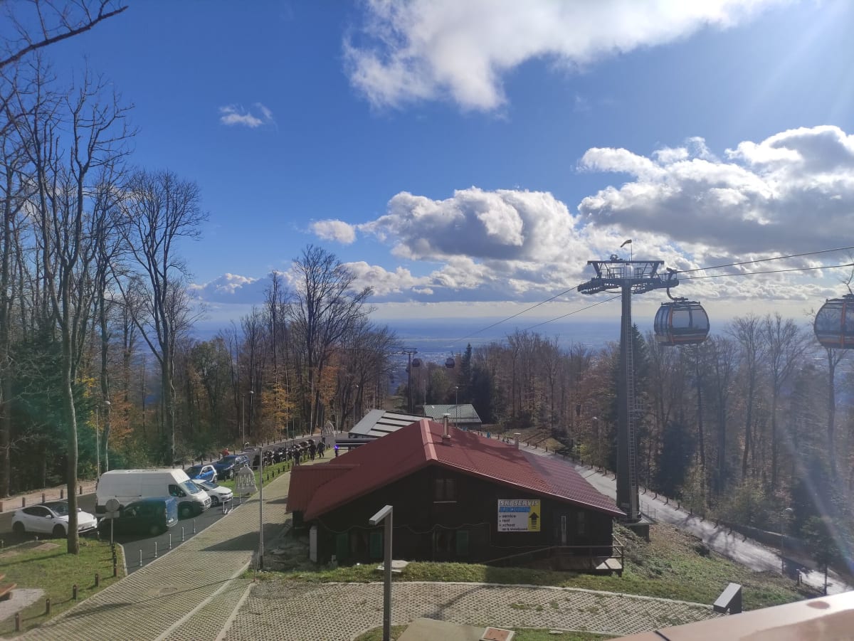 The Sljeme gondola cabins passing over a red-roofed ski-service chalet at the top station, the Pannonian plain hazy far below under a bright autumn sky