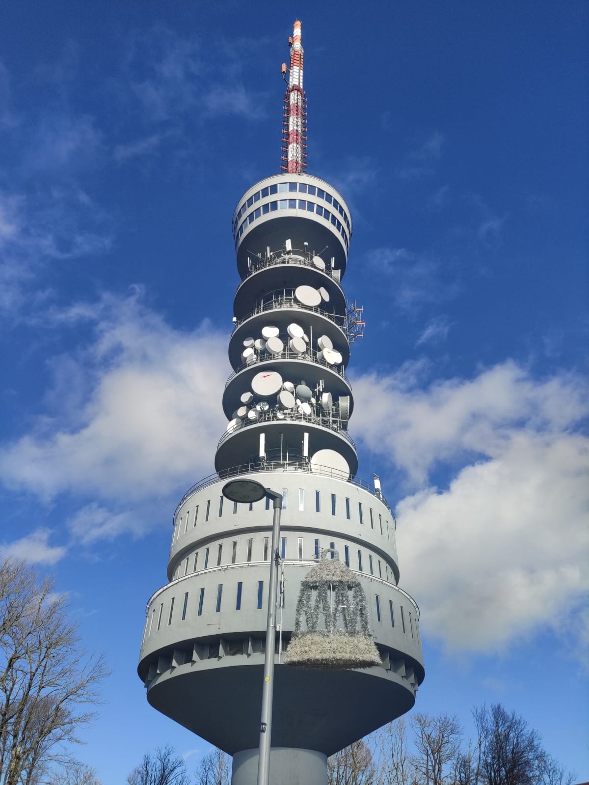 The Sljeme TV tower from directly below — tiered grey concrete rings hung with microwave dishes, topped by a red-and-white lattice antenna mast against a blue sky