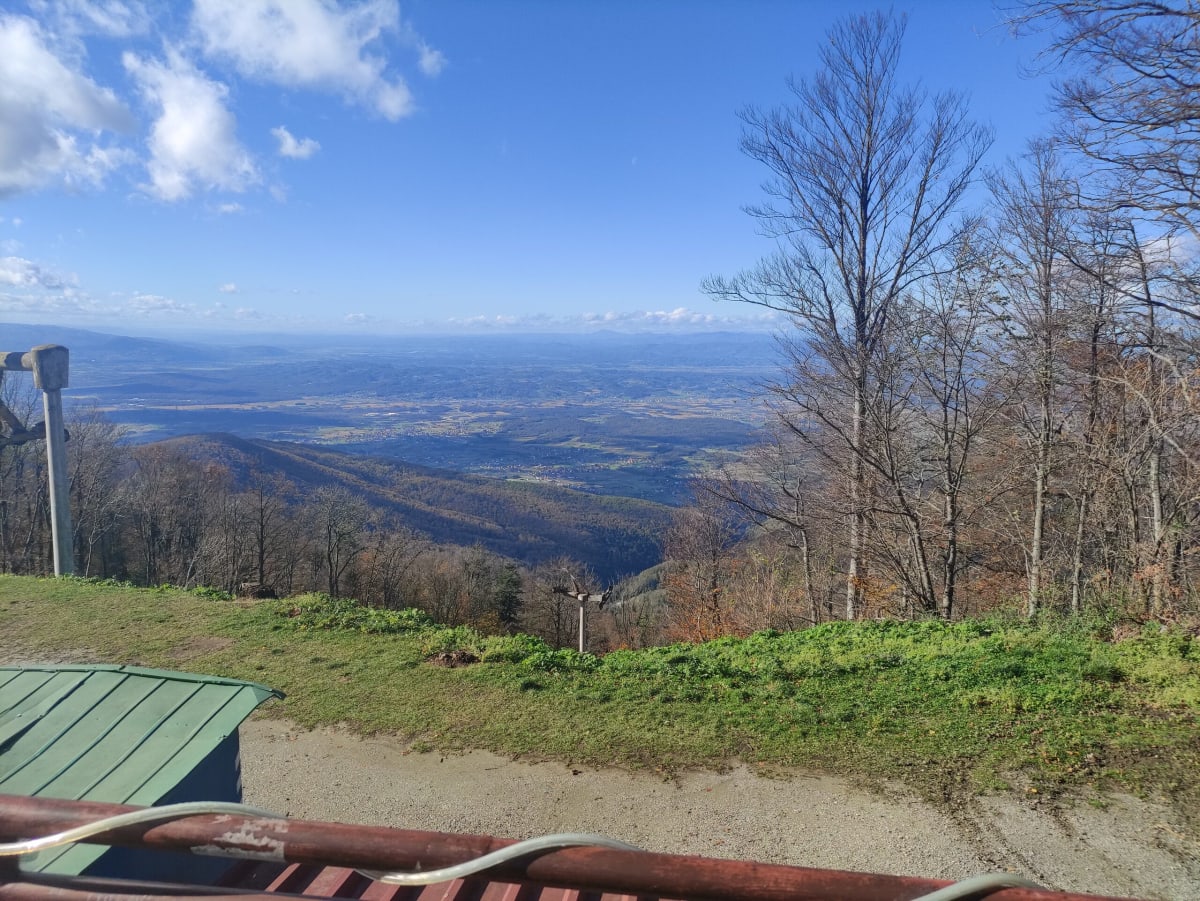 A viewpoint over the Zagorje hills from a bench beside an old ski drag-lift pylon, bare beech woods dropping away to the plain