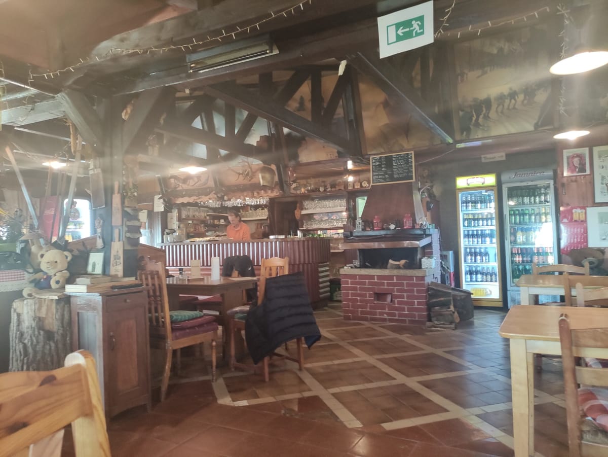 Inside a rustic mountain lodge — timber roof trusses, a corner bar, a big open brick fireplace, drinks fridges and old skis and photos on the walls