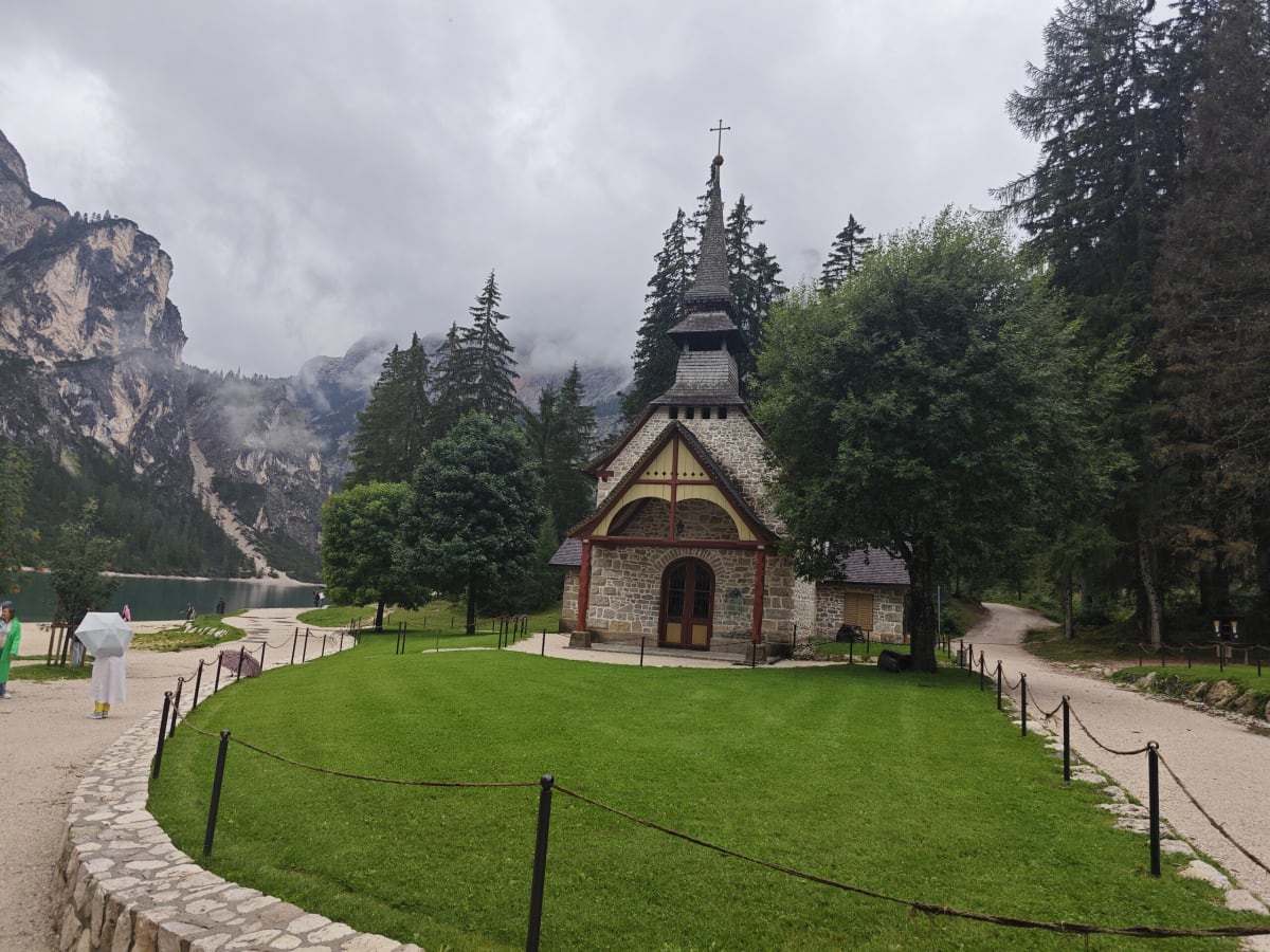 A small wooden-and-stone chapel with a steeple on a green lawn by Lago di Braies, steep Dolomite cliffs and low cloud behind it