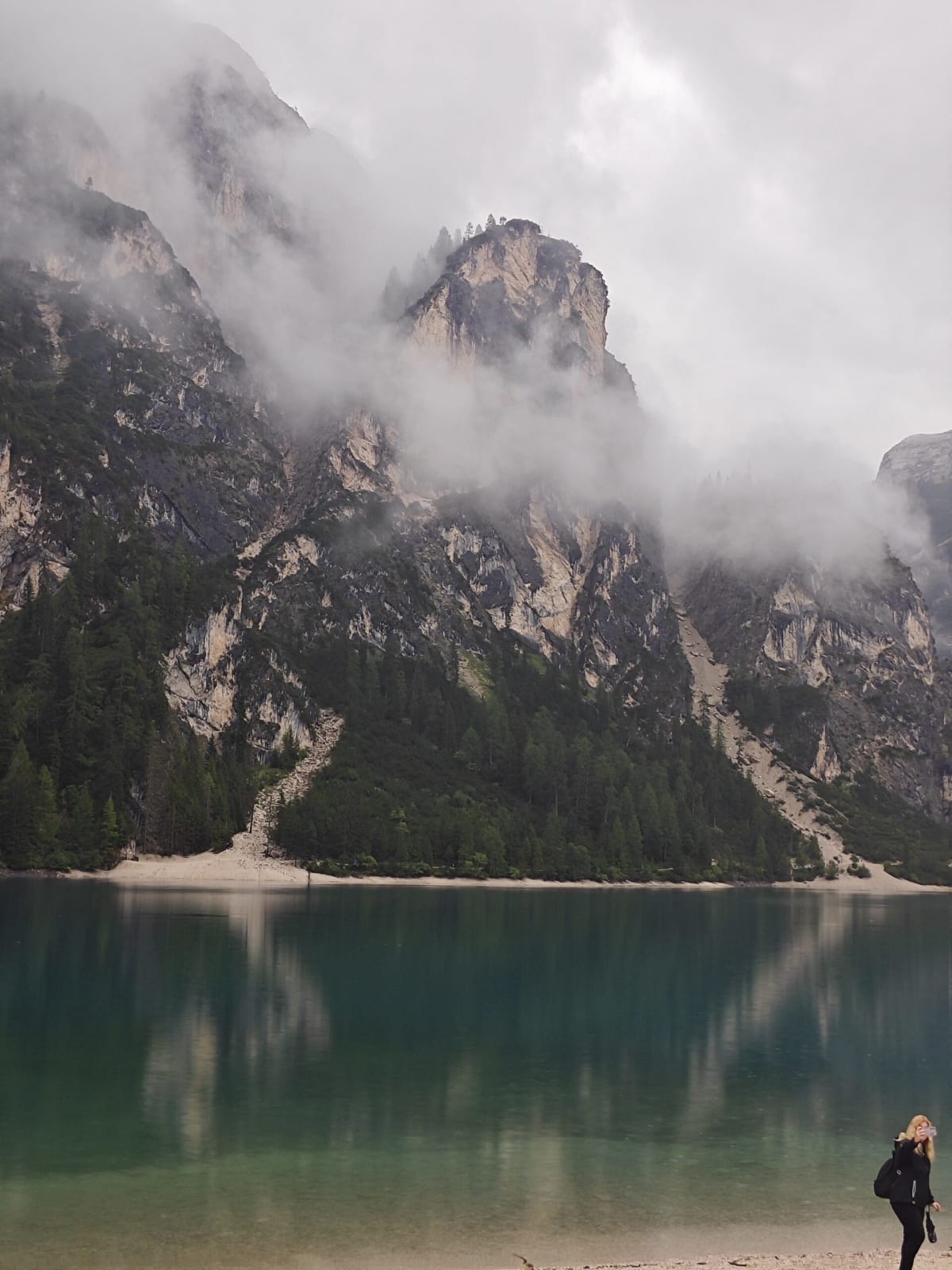 Mist drifting down a rocky Dolomite face above Lago di Braies, the still green water reflecting the cliffs and a visitor on the shore